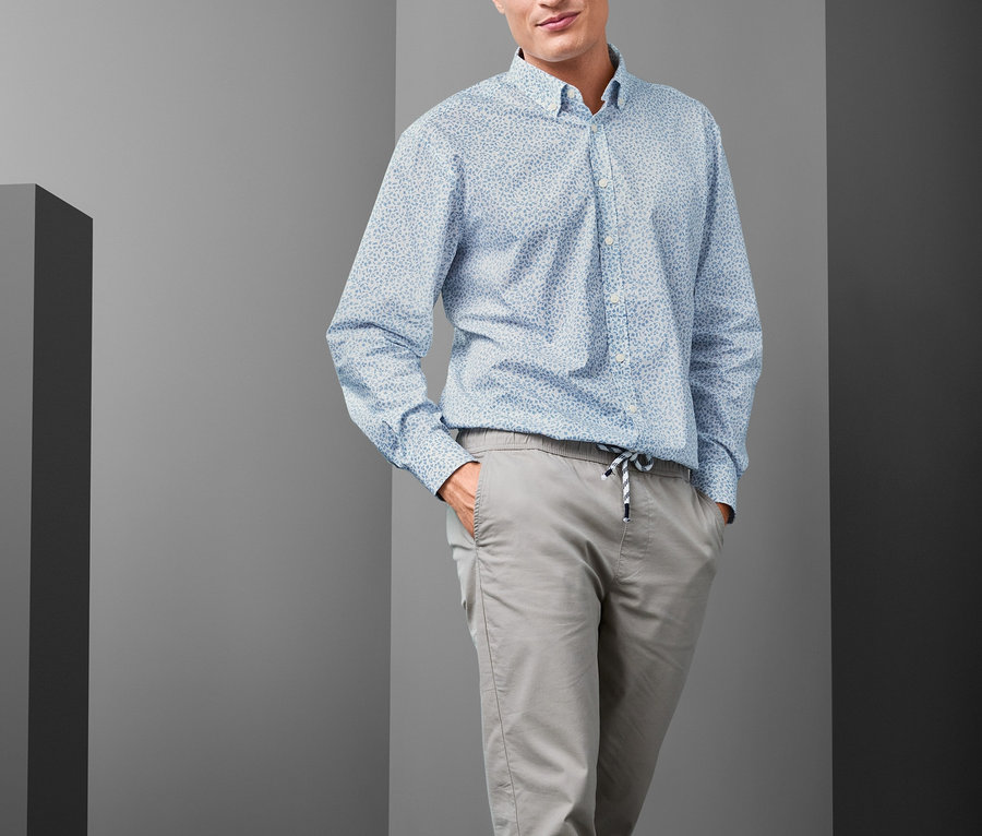 Muž pózuje a má na sebe svetlomodrú košeľu s golierom s gombíkmi Comfort Fit, sivé nohavice a modré tenisky.