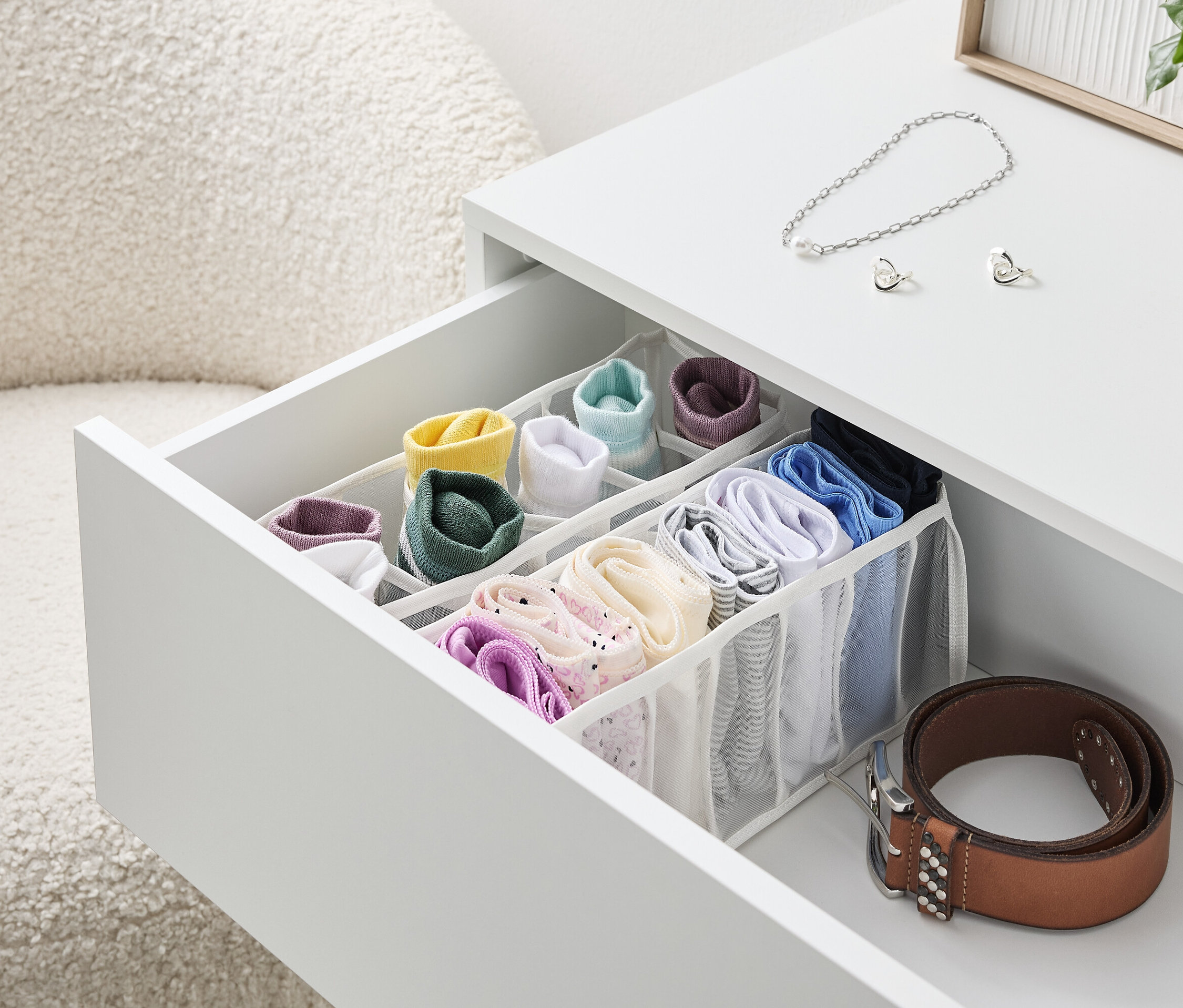 Otvorená zásuvka bieleho stolíka s organizérom plným farebných ponožiek a spodnej bielizne, vedľa leží hnedý opasok.
