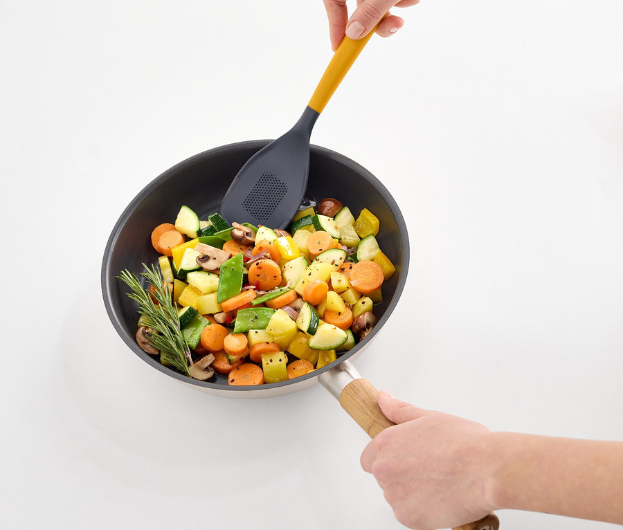 Ruky miešajú zeleninu na panvici žltou dierovanou obracačkou.