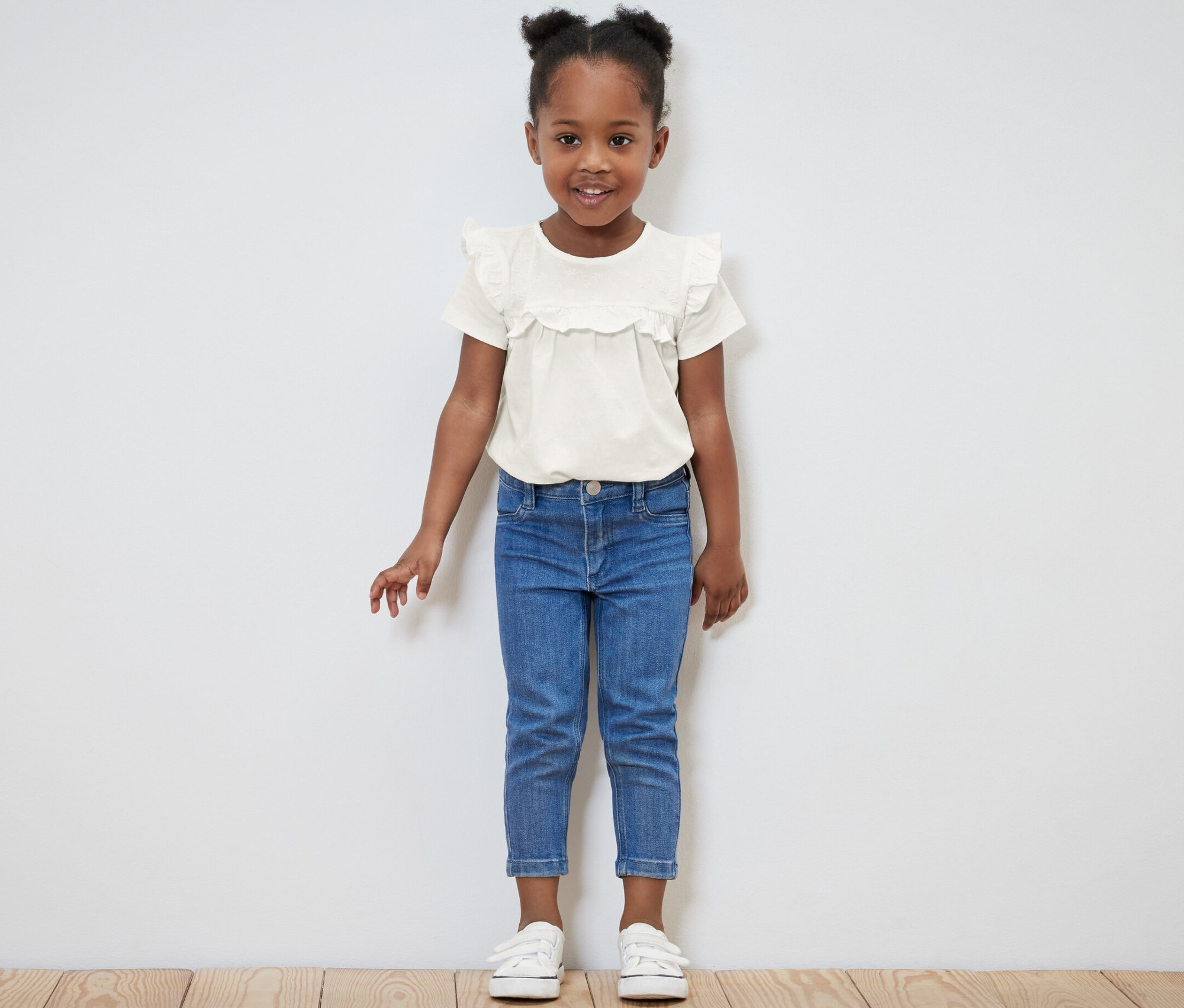 Usmievajúce sa dievčatko pózuje vo svetlom detskom tričku a modrých džínsoch.