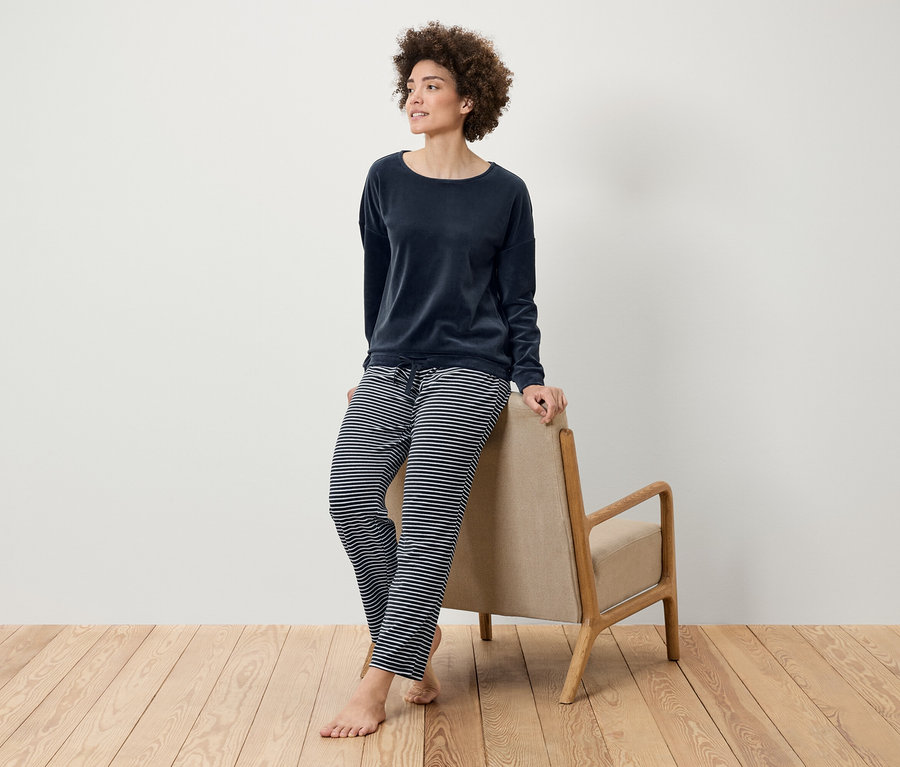 Žena s afro účesom pózuje v tmavomodrom zamatovom tope a pruhovaných nohaviciach, opierajúc sa o béžové kreslo na drevenej podlahe.