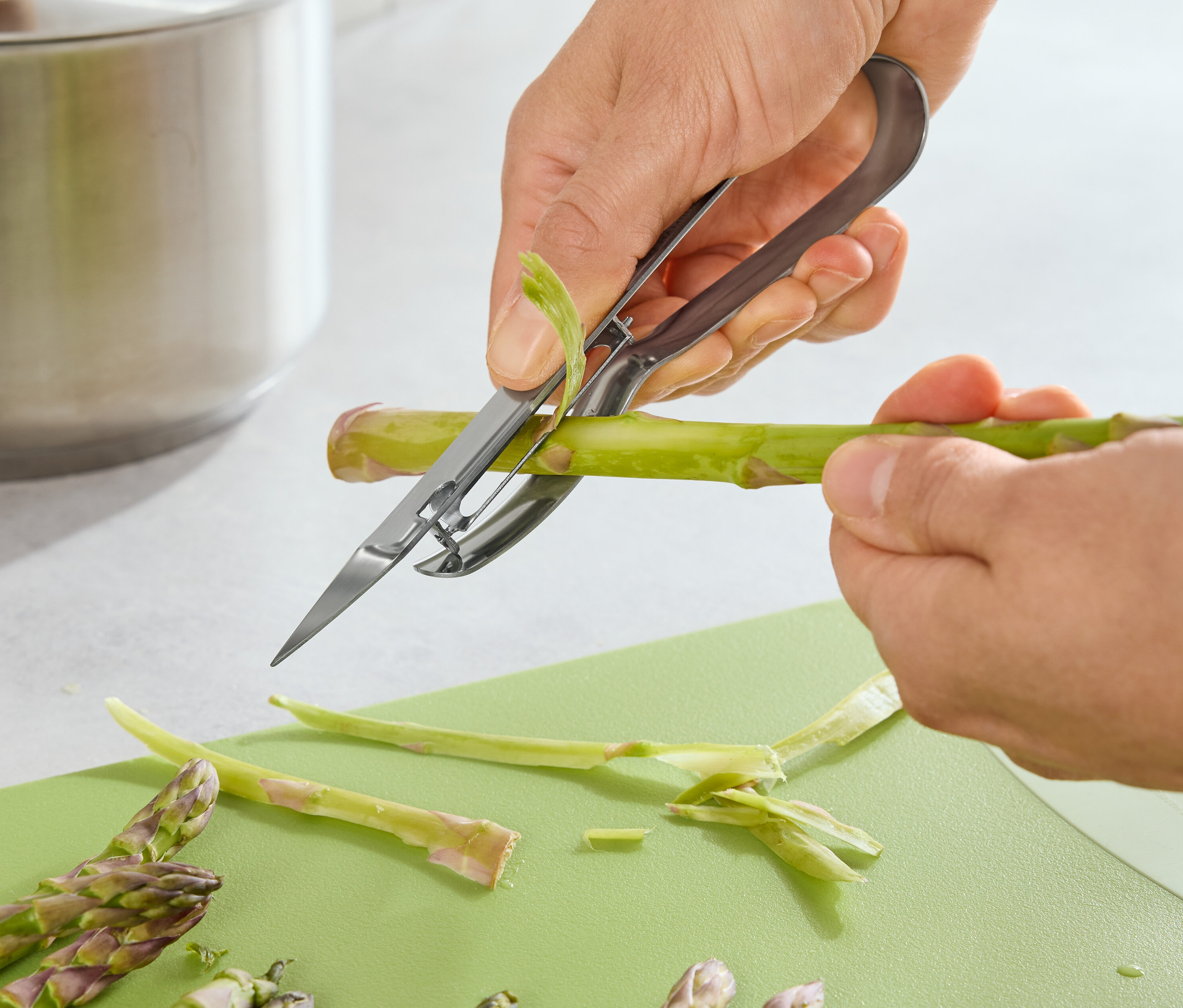 Osoba šúpe špargľu škrabkou na špargľu na zelenej doske na krájanie.
