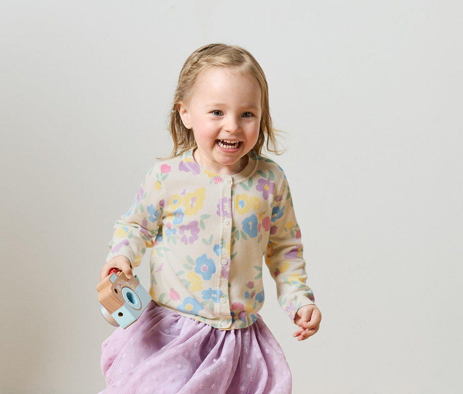 Usmievavé blond dievča so zapletenými vrkočmi v kvetinovom kardigane a fialovej sukni drží hračkársky fotoaparát.