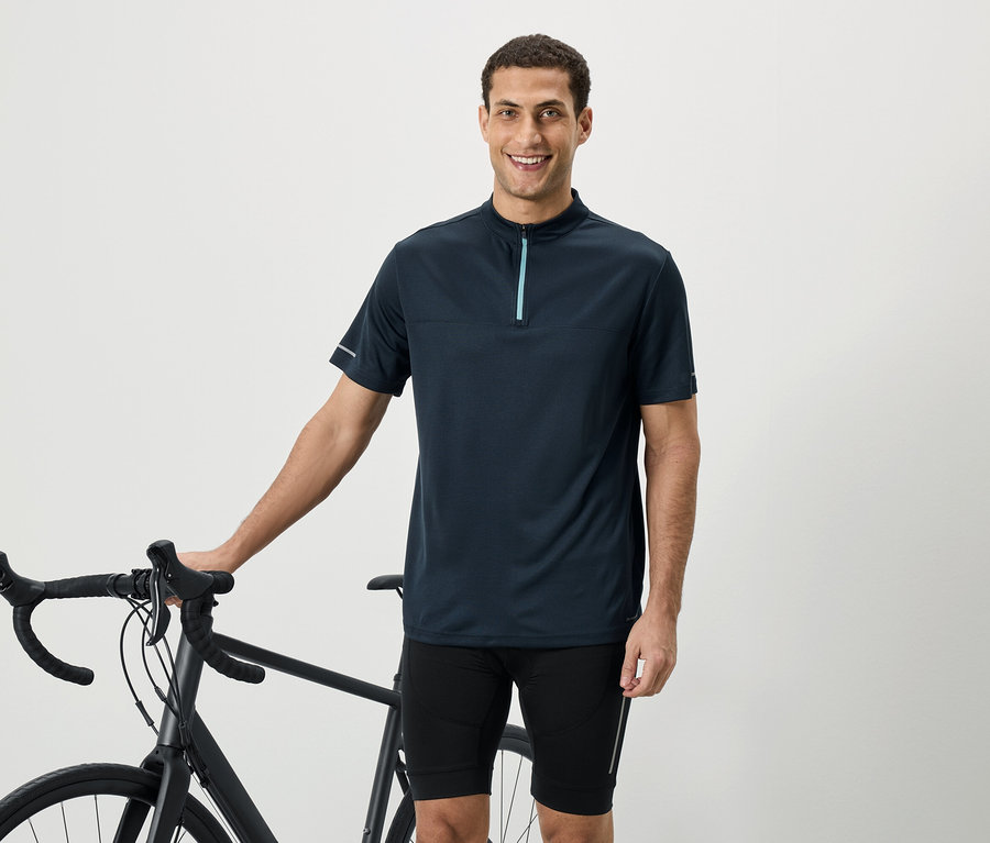 Usmievavý muž v tmavomodrej košeli s krátkym rukávom so zipsom stojí vedľa bicykla.
