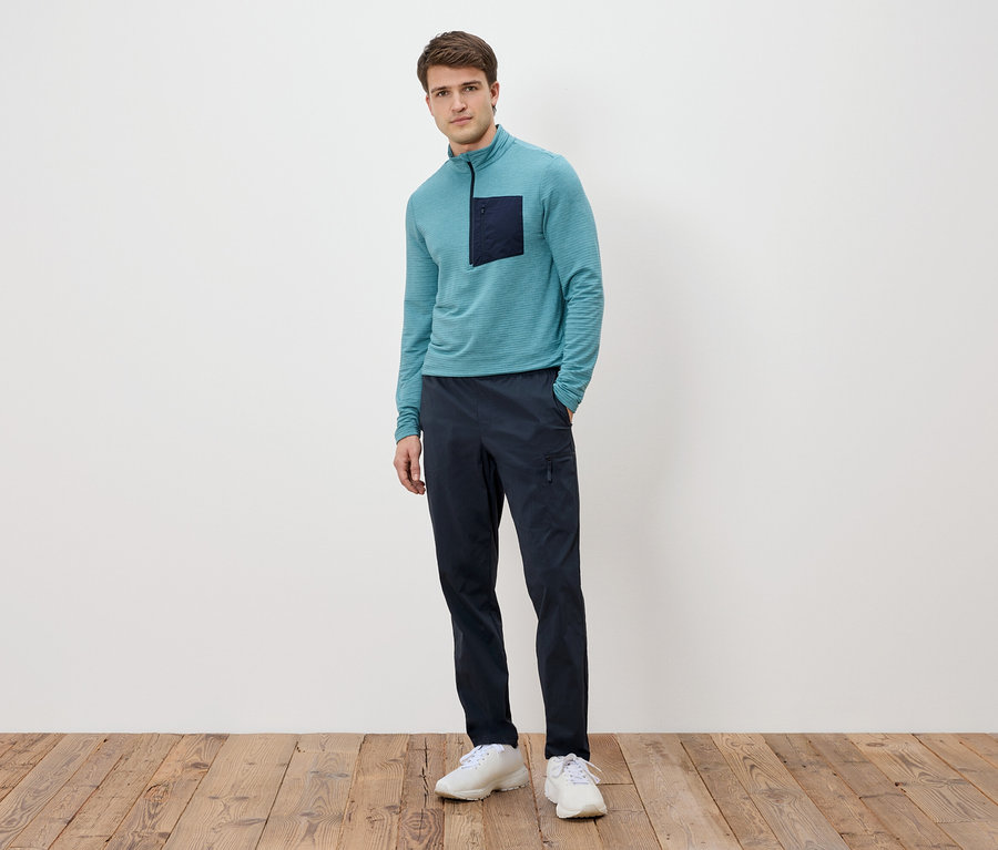 Muž stojí pred bielou stenou, má na sebe tyrkysové tričko s dlhým rukávom s tmavým vreckom, tmavomodré nohavice a biele tenisky.