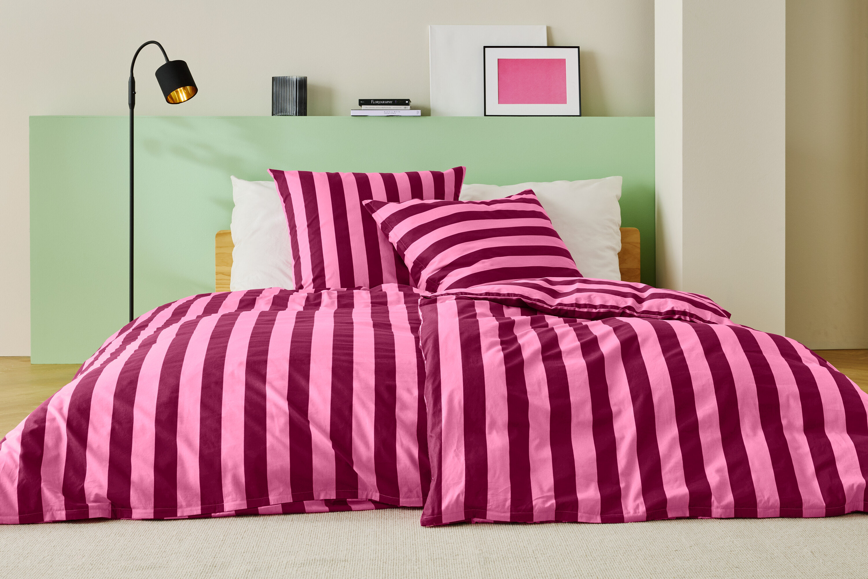 Posteľ s ružovo-vínovočervenými pruhovanými obliečkami, dvoma vankúšmi a stojacou lampou.
