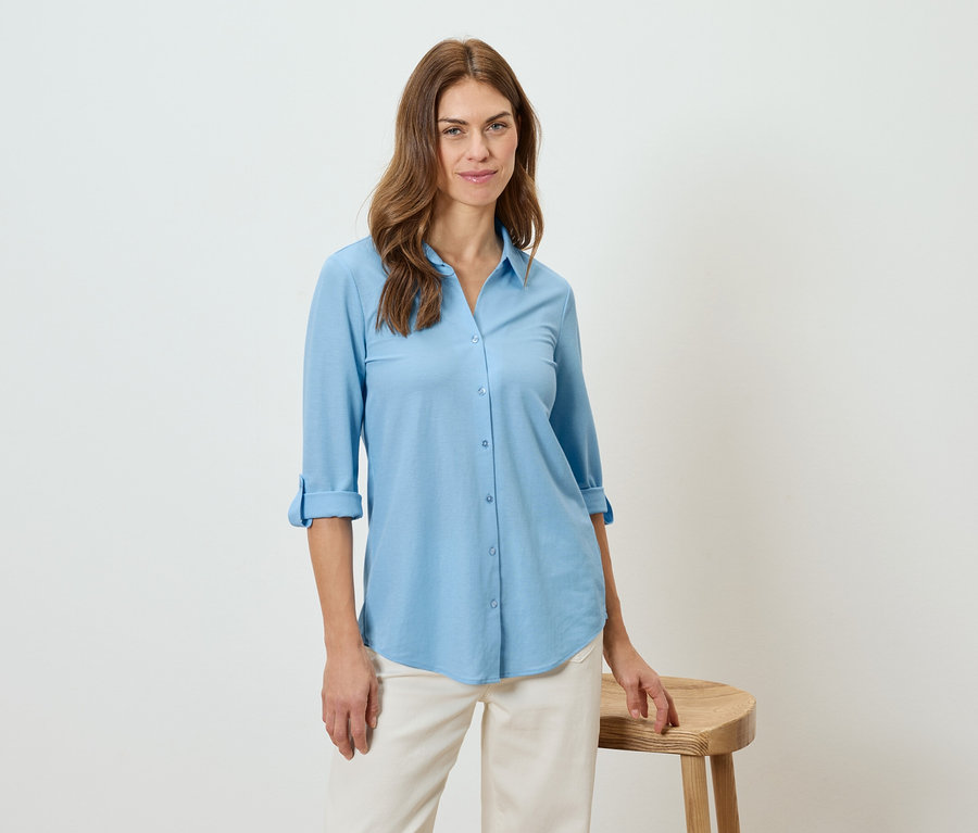 Žena má na sebe svetlomodrú košeľu a béžové nohavice, opiera sa o stoličku.