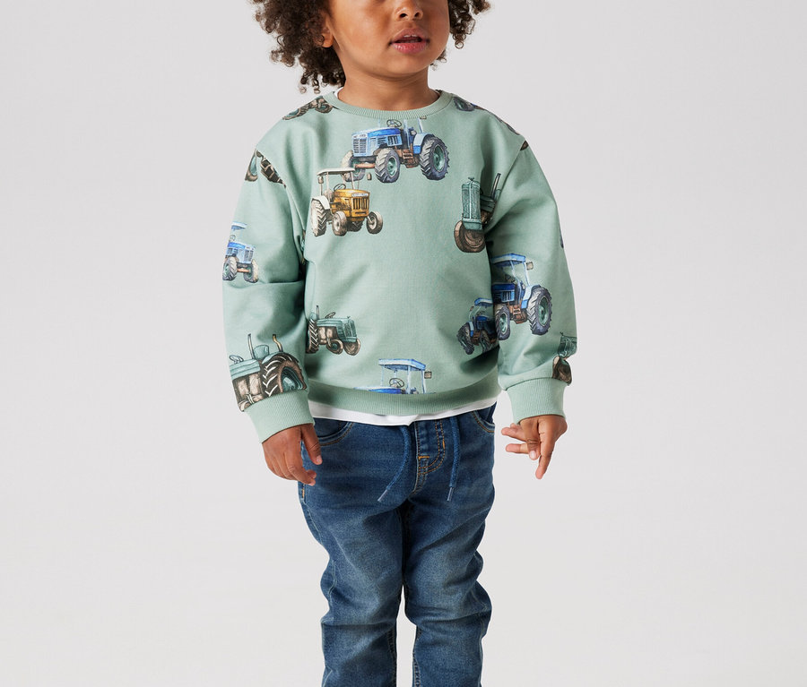 Malý chlapec s kučeravými vlasmi stojí v mätovo zelenom svetri s traktormi a džínsoch.