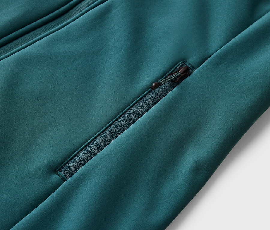 Detailný záber smaragdovo zelené bundy s vreckom na zips a ďalším zipsom v ľavom hornom rohu.