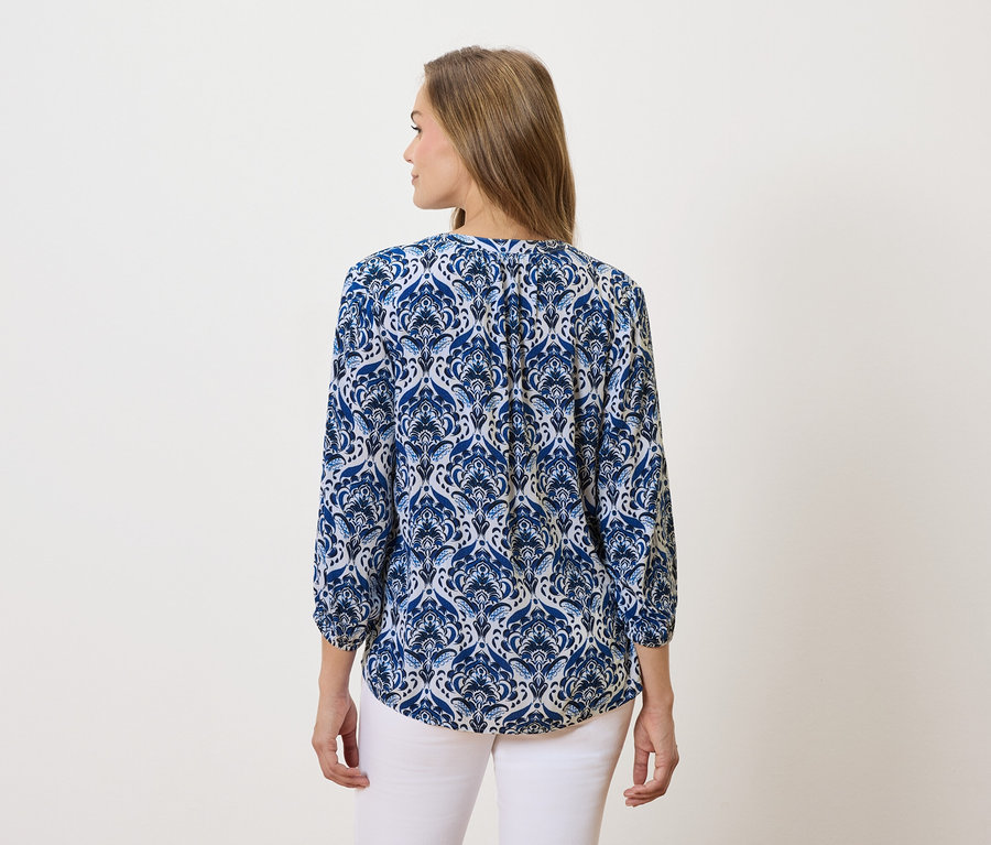 Žena s dlhými hnedými vlasmi má na sebe modro-bielu vzorovanú blúzku a biele nohavice.