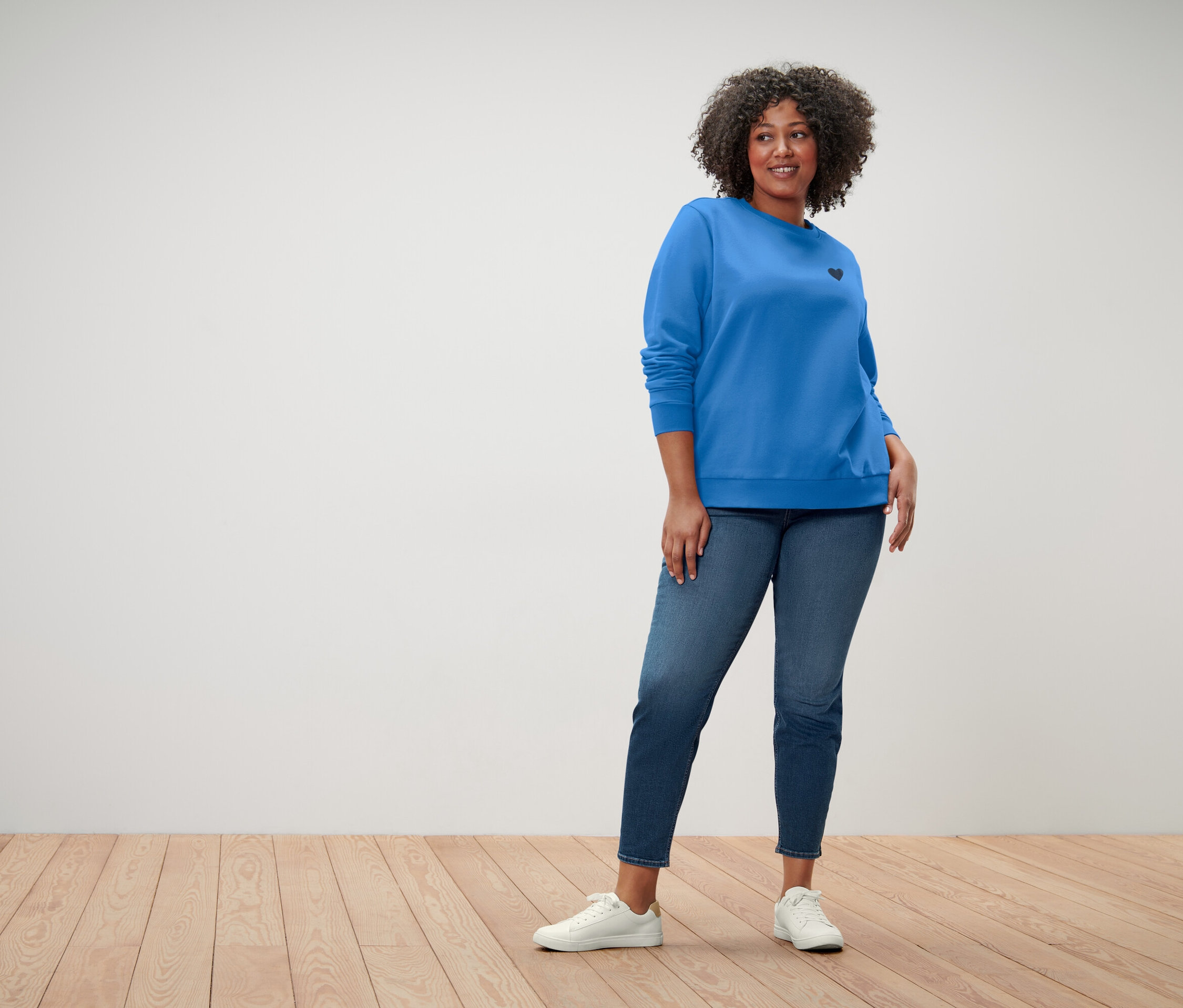 Žena s kučeravými vlasmi má na sebe modrú mikinu s malým čiernym srdcom, modré džínsy a biele tenisky.