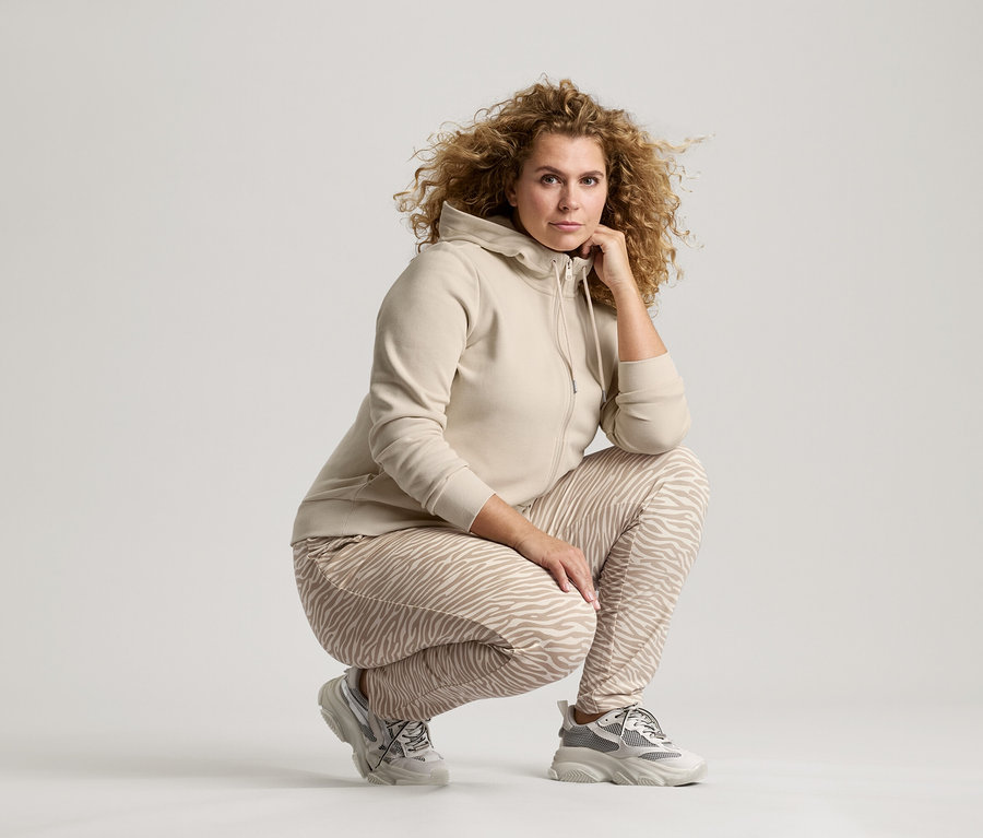 Žena s kučeravými vlasmi v béžovej bunde, nohaviciach so vzorom zebry a teniskách sa krčí s rukou na brade.