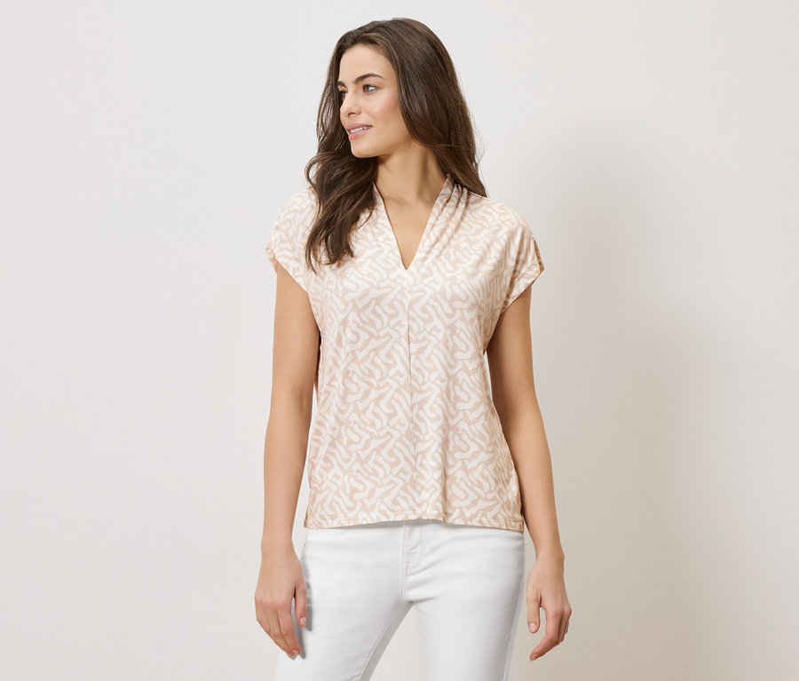 Žena má na sebe svetlohnedú vzorovanú blúzku s výstrihom do V a biele nohavice.