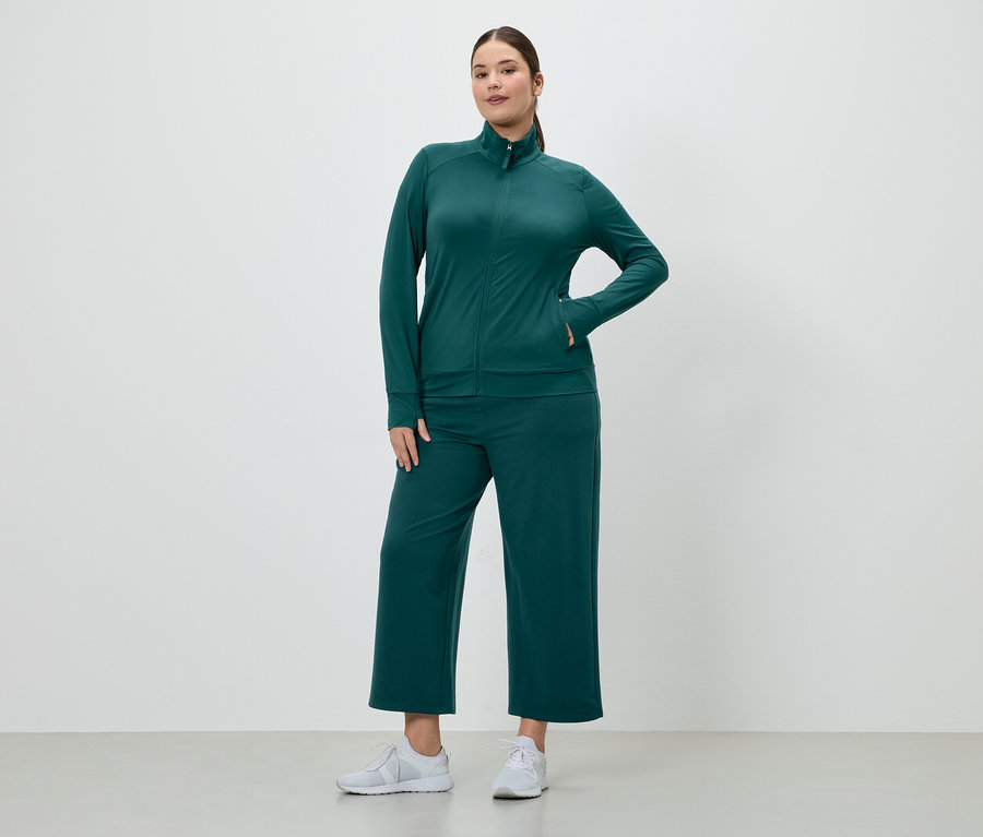Žena v smaragdovozelenej teplákovej súprave so zapínacou bundou a culottes nohavicami a bielych teniskách.