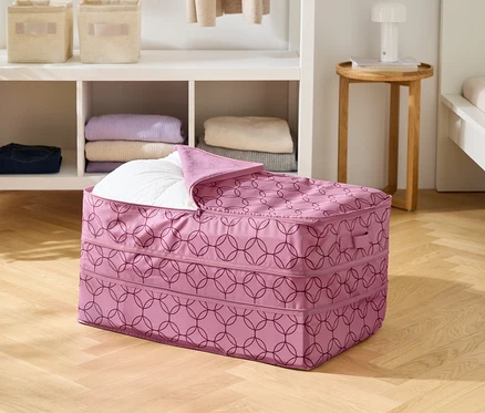 Na drevenej podlahe stojí ružová úložná taška s geometrickým vzorom, čiastočne vyplnená bielou prikrývkou.