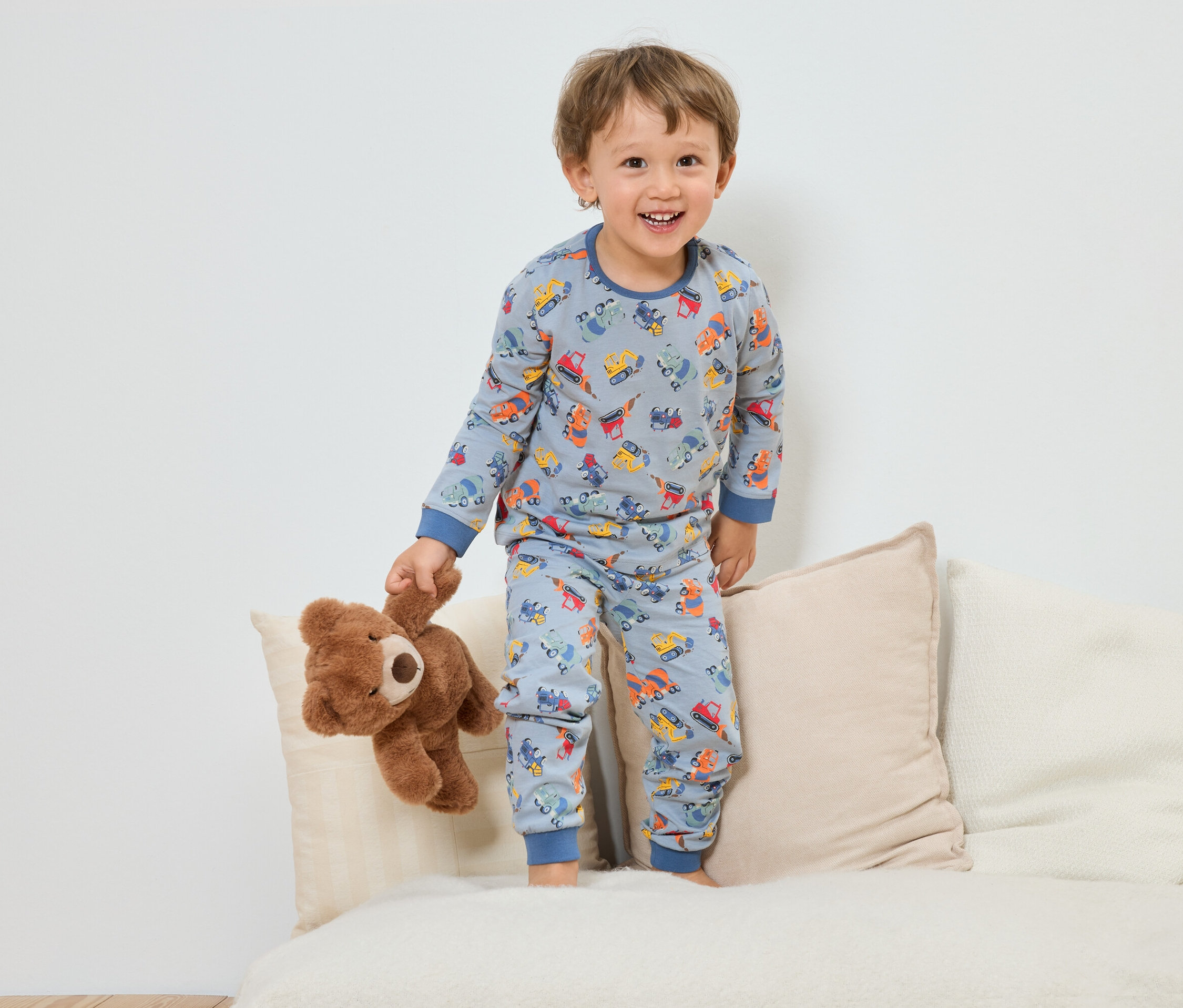 Usmievavý chlapec v modrom pyžame so vzorom stavebných vozidiel stojí s plyšovým medvedíkom na pohovke.