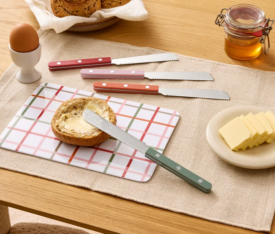 Na stole leží natretá žemľa, štyri nože, maslo, med, vajce a chlieb.