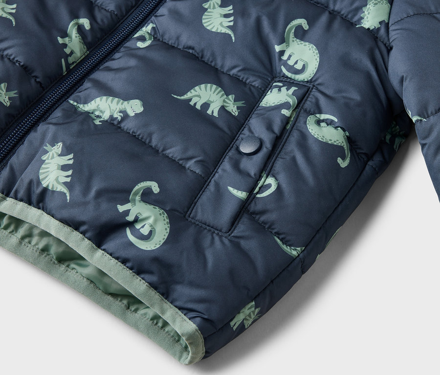 Detailný záber na tmavomodrú prešívanú bundu so svetlozeleným vzorom dinosaurov, zipsom a vreckom so zapínaním na patentku.
