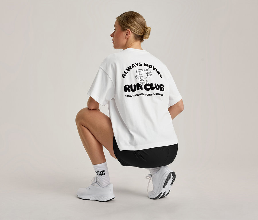 Žena s blond drdolom čupí, videná zozadu. Má na sebe biele, oversized športové tričko s čiernou grafikou 'ALWAYS MOVING RUN CLUB' na chrbte, čierne šortky, biele ponožky a biele športové topánky.