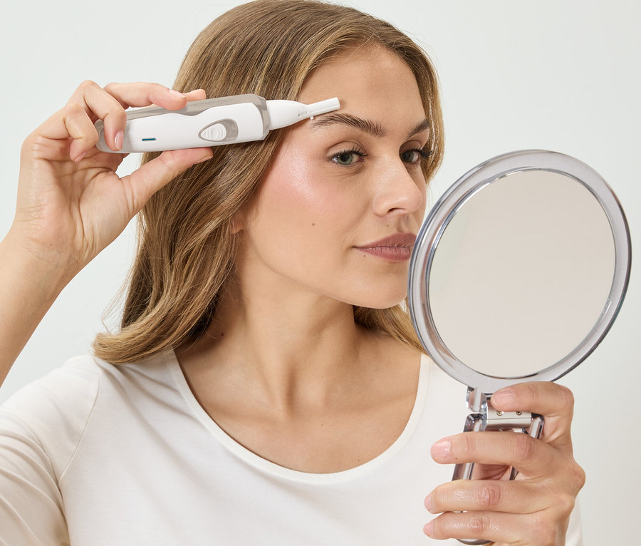 Žena si upravuje obočie bielym epilátorom, pričom sa pozerá do okrúhleho zrkadla.