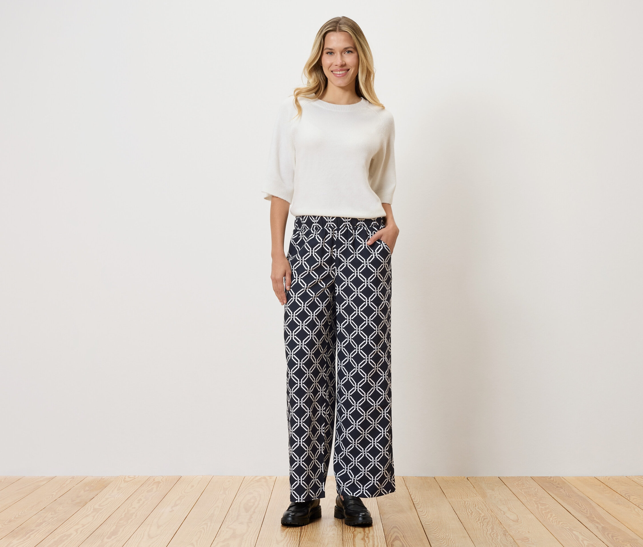 Žena v bielom tope a vzorovaných nohaviciach stojaca na drevenej podlahe pred stenou.