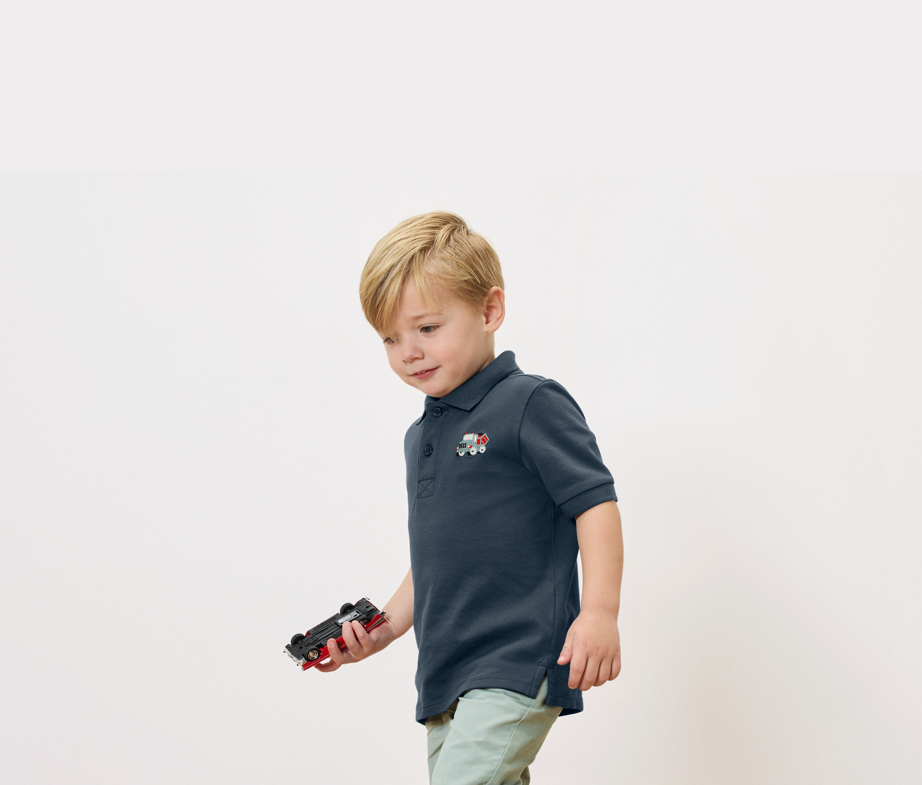 Blond chlapec drží v ruke autíčko a má na sebe tmavomodré polokošele s malou aplikáciou vlaku.