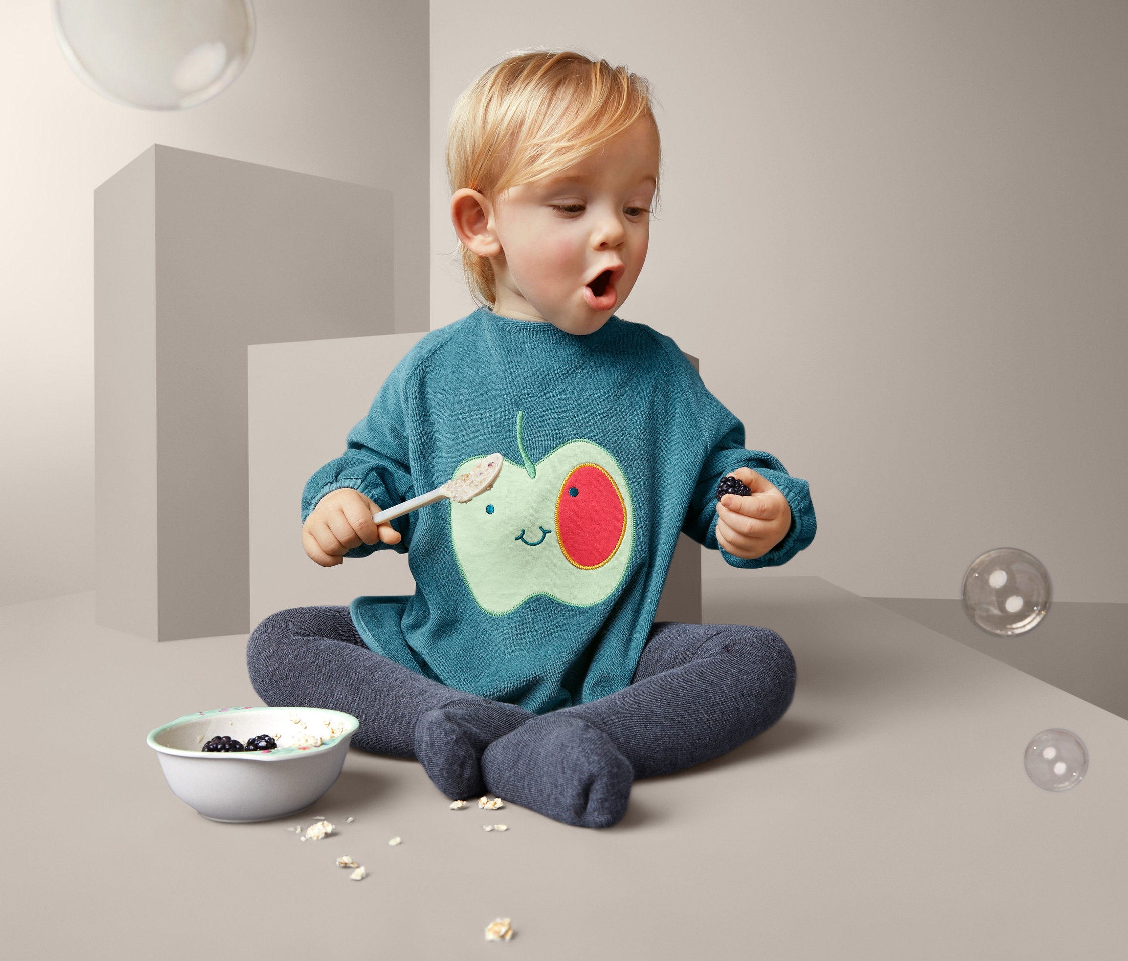 Batoľa sedí s miskou a lyžicou, má na sebe podbradník s jablkom.