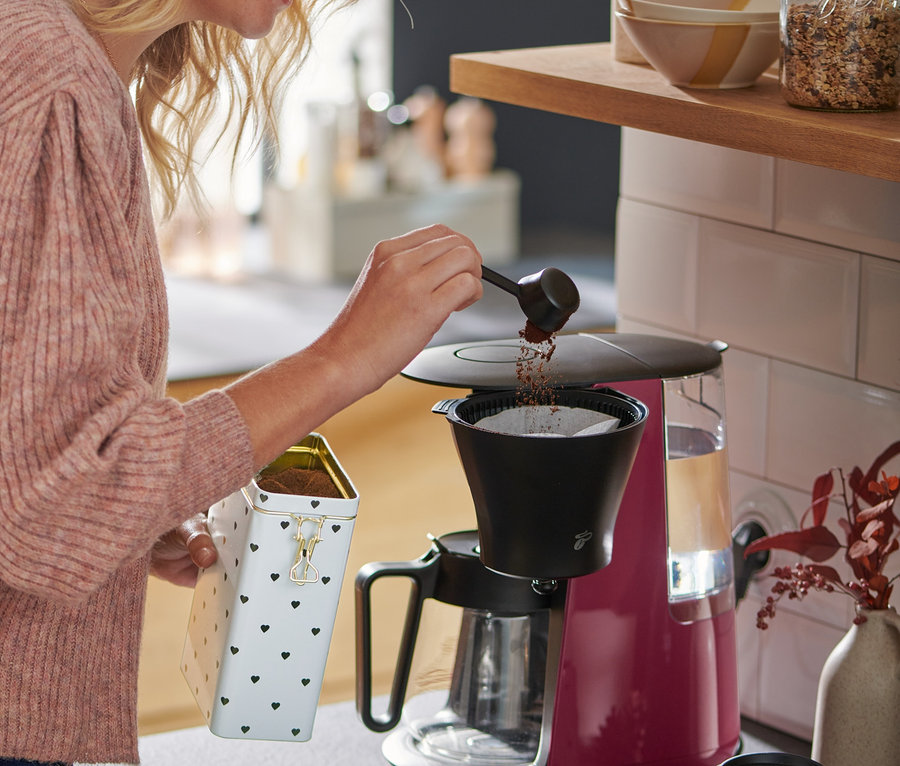 Žena s blond vlasmi sype kávu do malinového kávovaru na prekvapkávanú kávu »Let's Brew«.