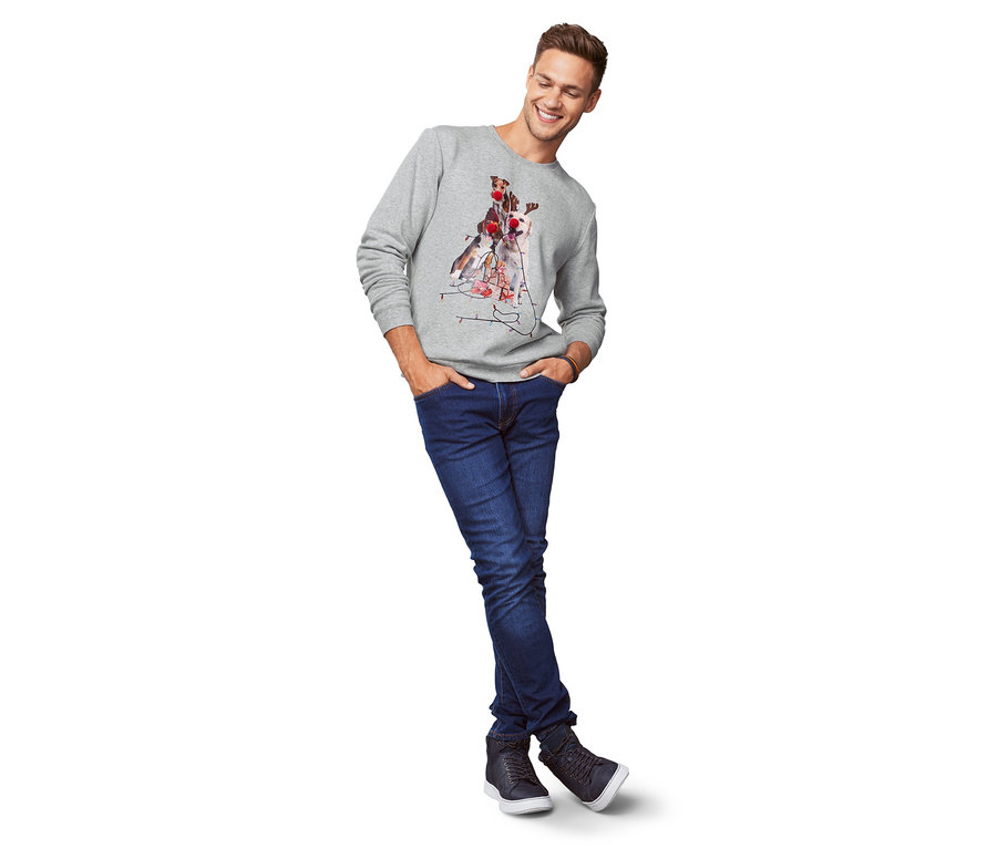 Muž v šedej mikine s potlačou psov a džínsoch stojí s rukami vo vreckách.