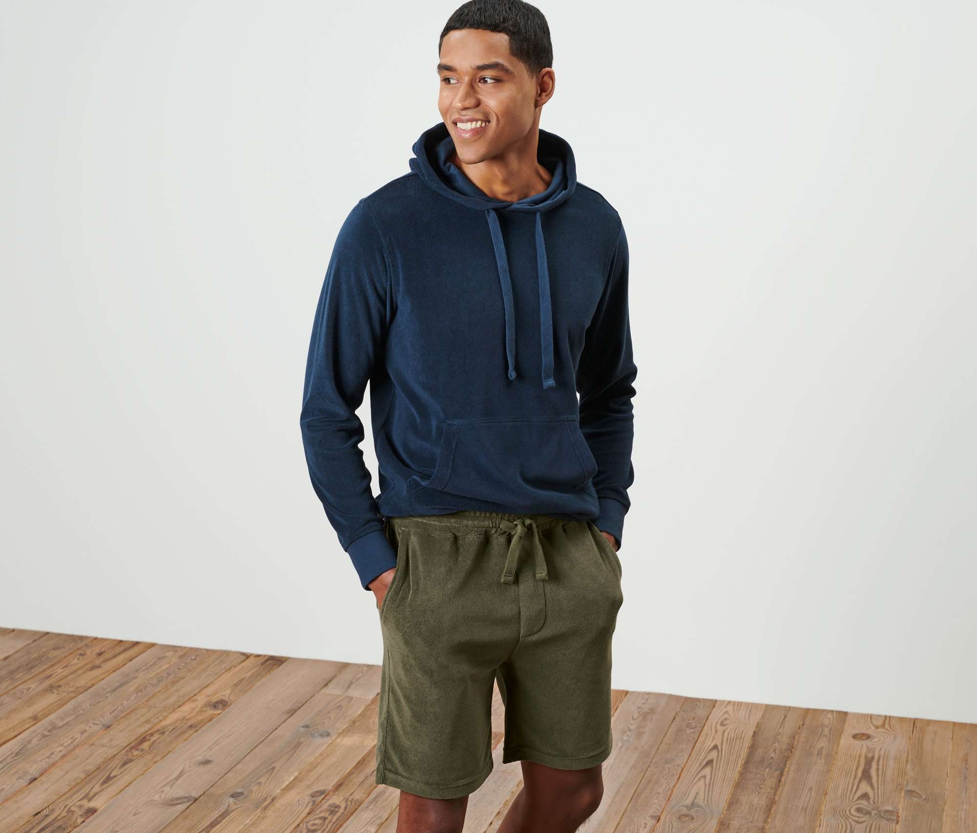 Muž s úsmevom stojí a má na sebe modrú mikinu s kapucňou a zelené froté šortky.