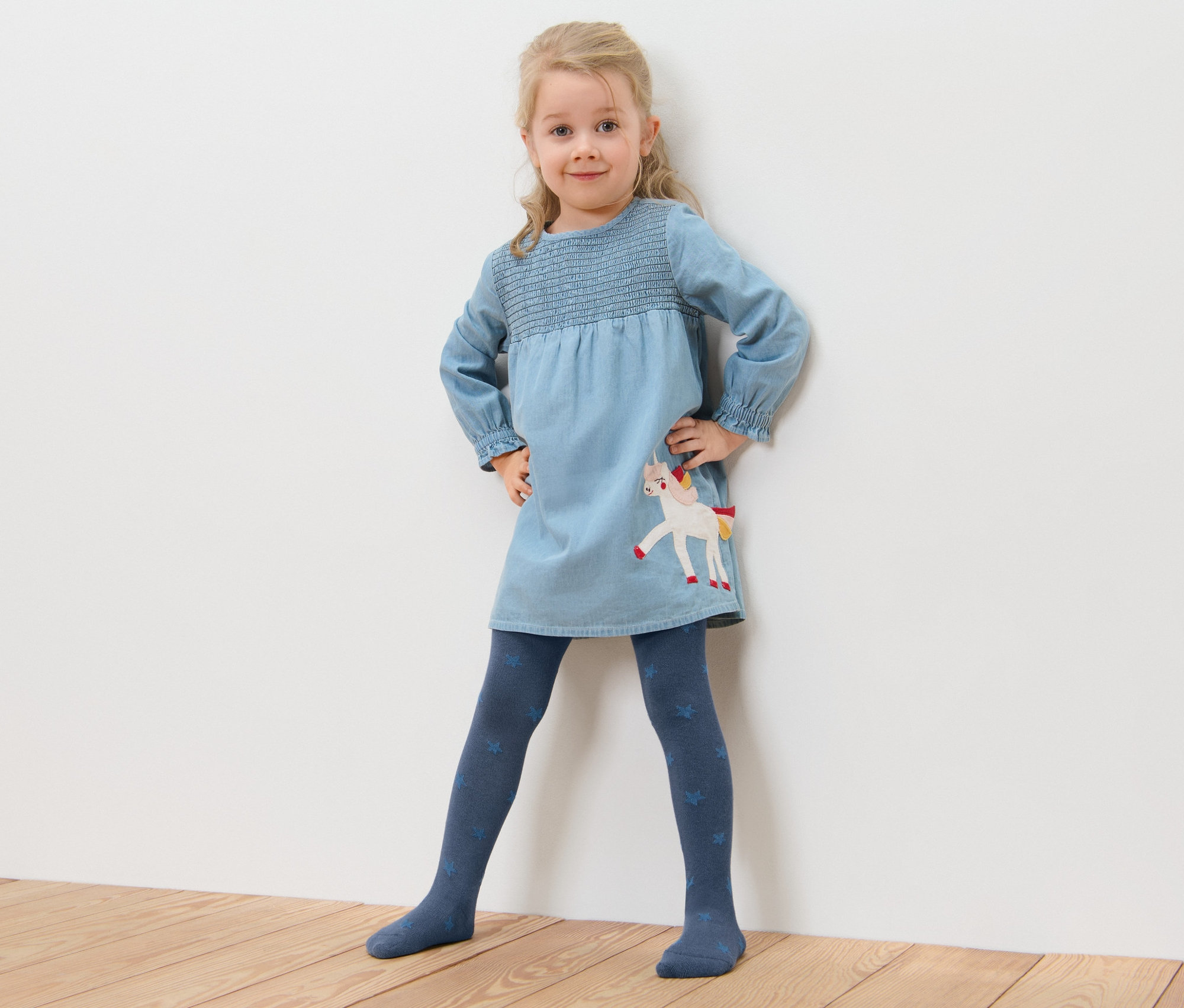 Dievčatko pózuje v modrých šatách s jednorožcom a bavlnených pančuchových nohaviciach s hviezdičkami (5 ks).