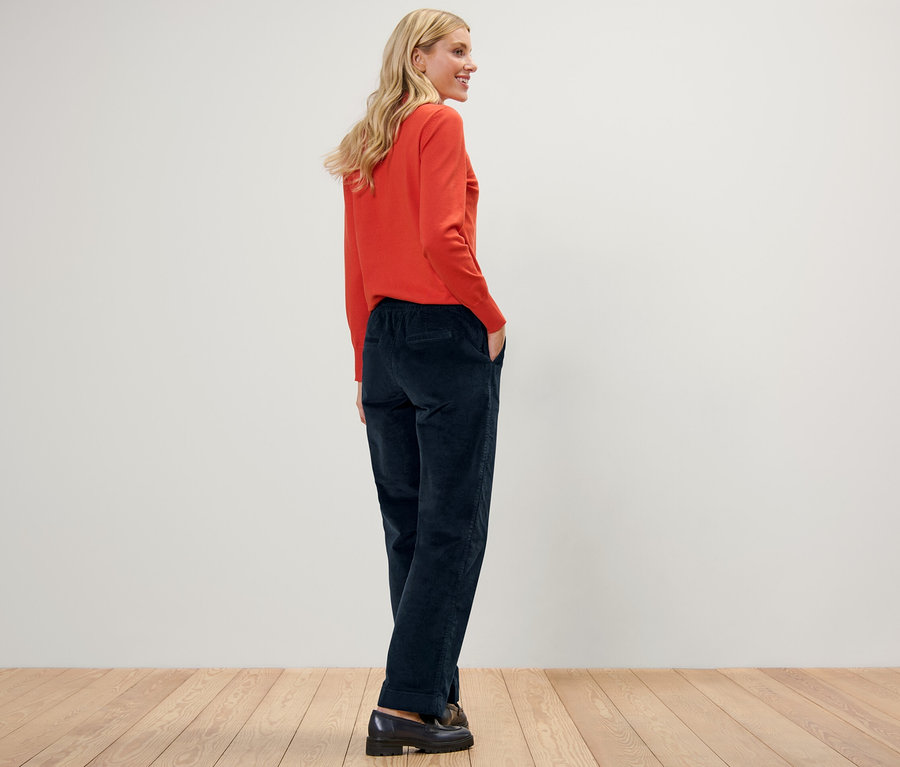 Žena pózuje v menčestrových pohodlných nohaviciach a oranžovom svetri.