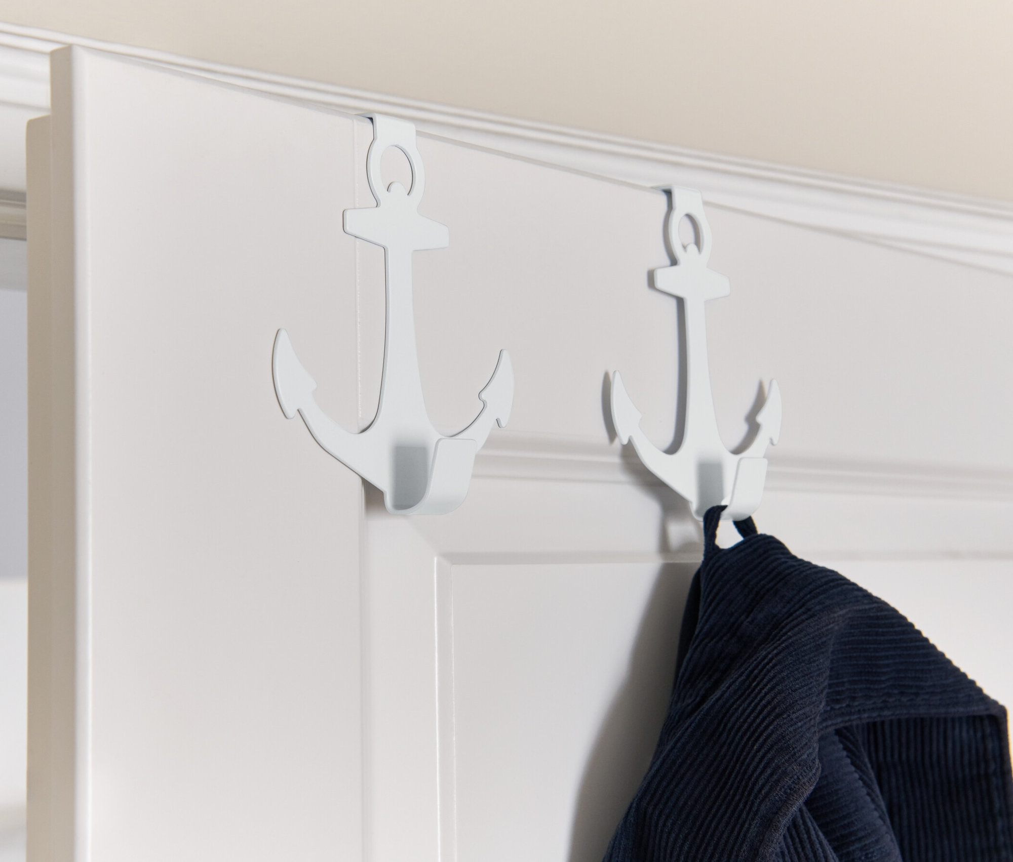 Dva biele háčiky na dvere v tvare kotvy visia na bielych dverách, na jednom z nich visí modré oblečenie.