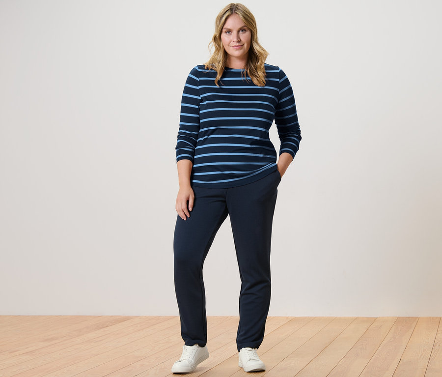 Žena stojí a má na sebe tmavomodré tepláky a prúžkované tričko s dlhými rukávmi.
