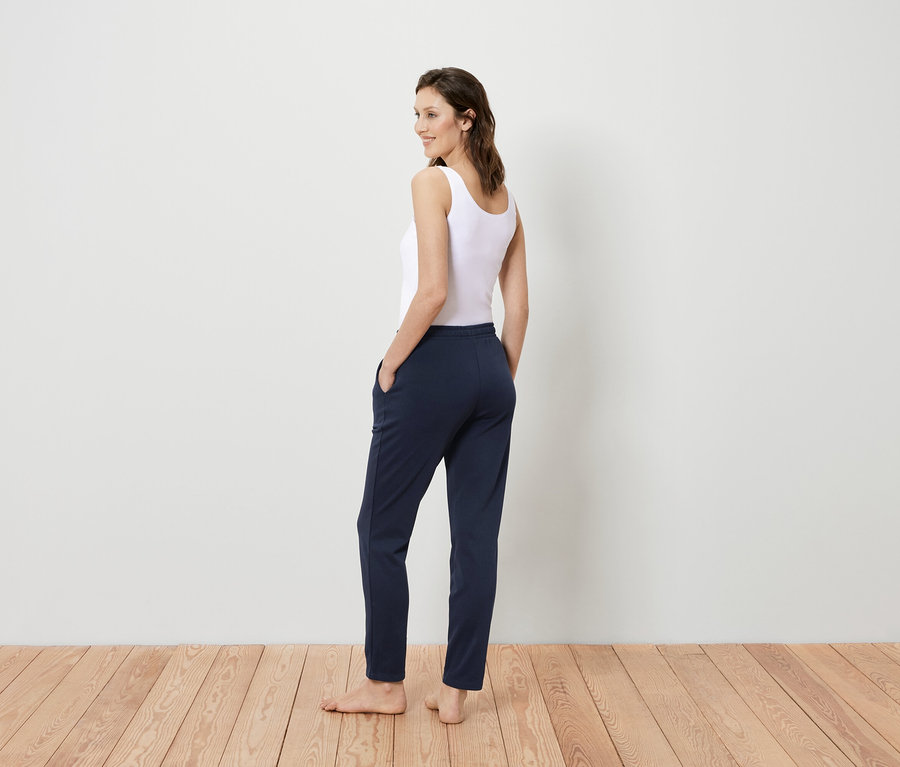 Žena v bielom tielku a tmavomodrých nohaviciach stojí chrbtom k divákovi a pozerá sa cez plece.