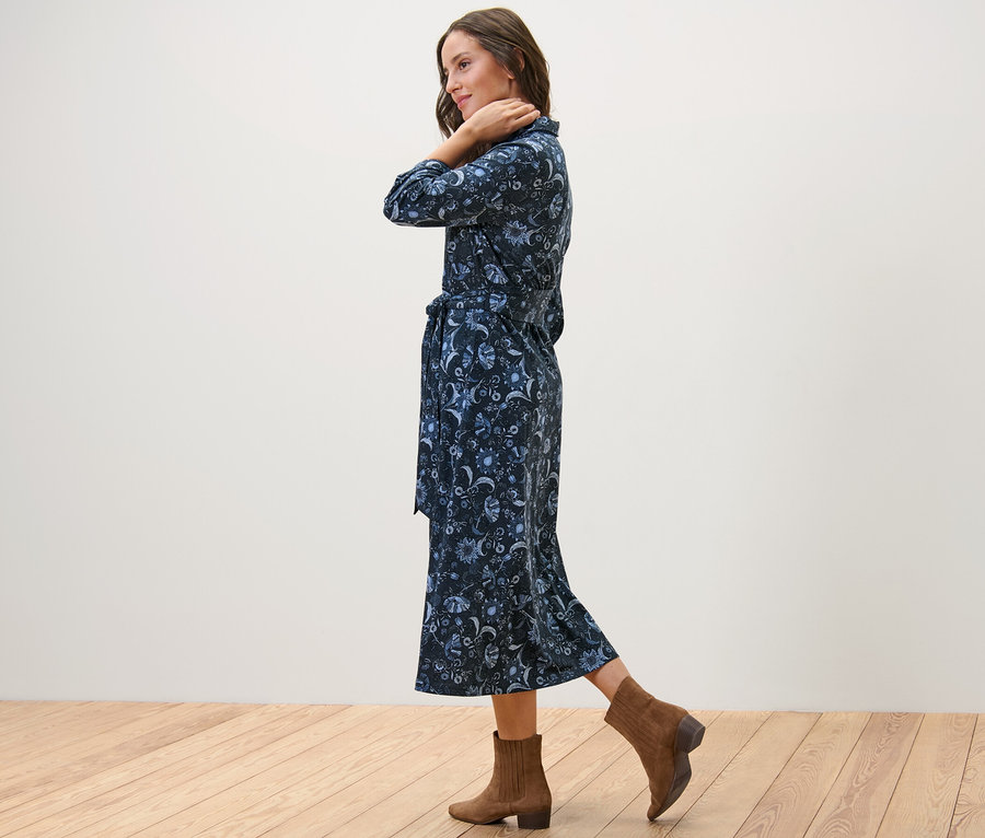 Žena pózuje bokom v džersejových šatách s potlačou a hnedých členkových topánkach.