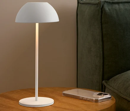 Biela lampa stojí na drevenom stolíku vedľa mobilného telefónu a zelenej pohovky.