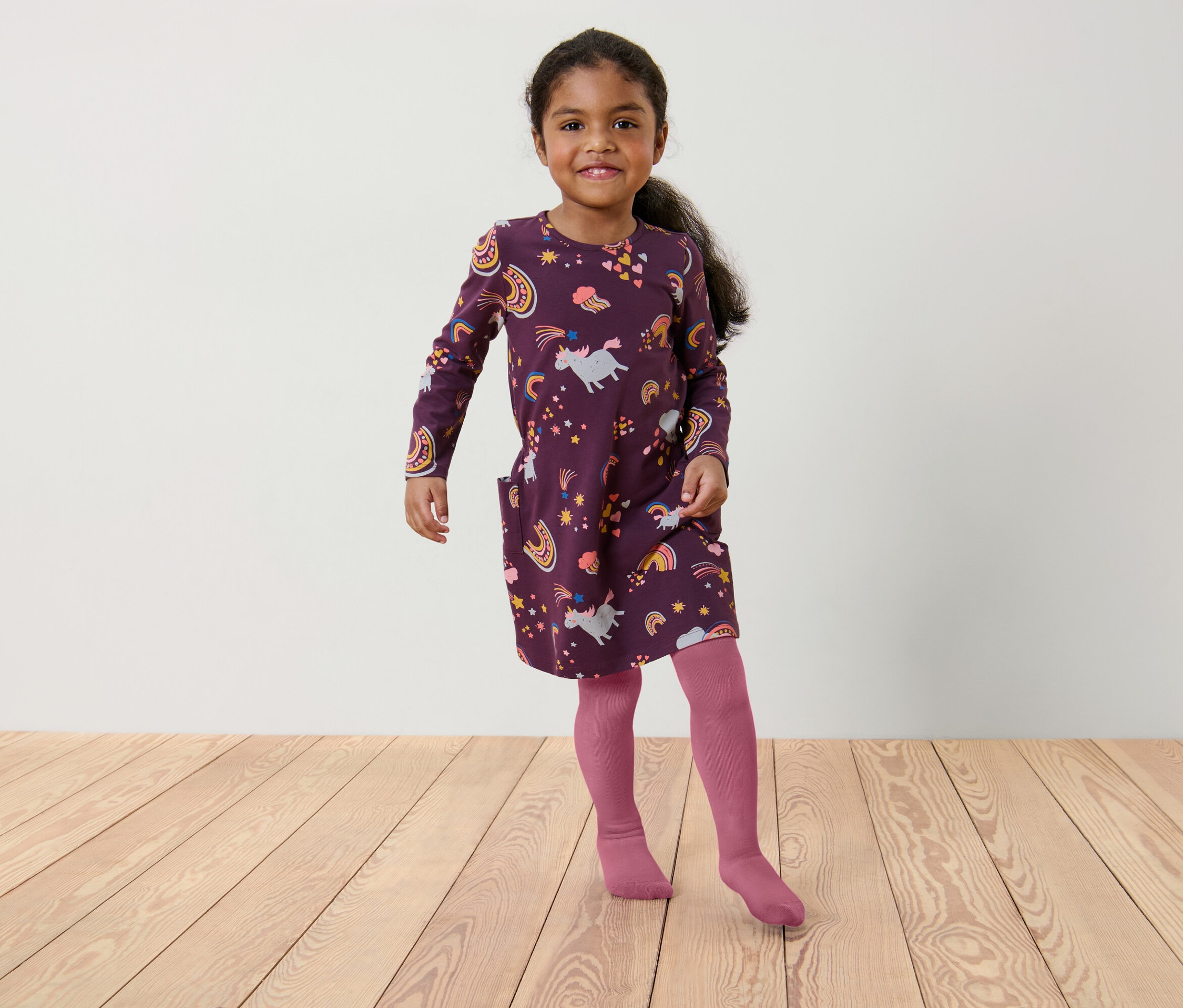 Dievča v bordových šatách s potlačou jednorožcov a dúhy a ružových pančuchách stojí na drevenej podlahe.