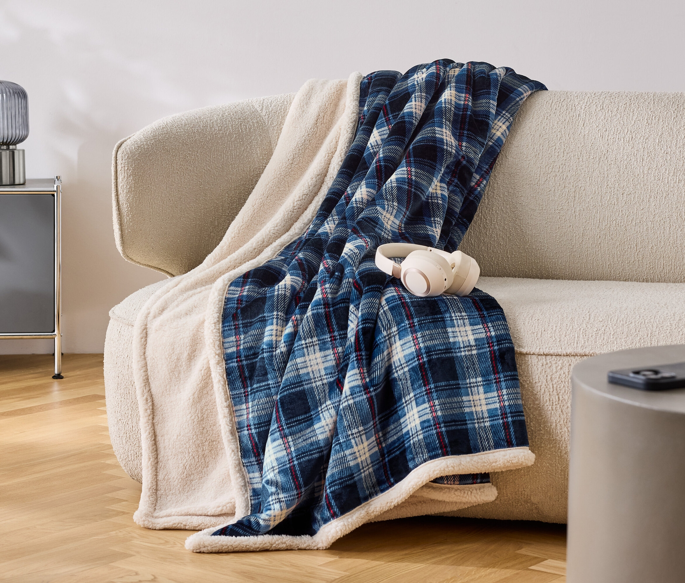 Na béžovej pohovke leží modro-biela kockovaná deka s bielym lemom. Na deke sú položené biele slúchadlá.