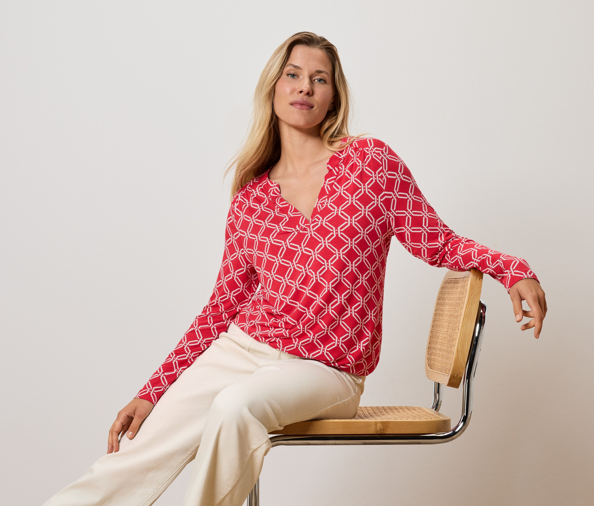 Blondínka sedí na stoličke, má na sebe červeno-biely top a krémové nohavice.