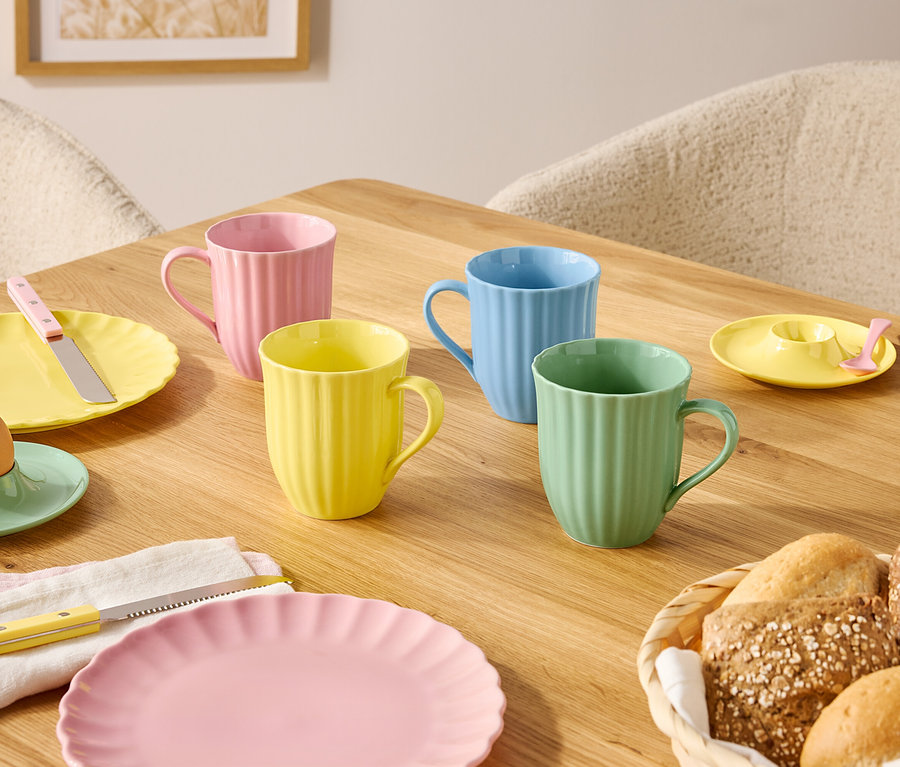 Na drevenom stole sú prestreté taniere a hrnčeky v pastelových farbách a košík s pečivom.