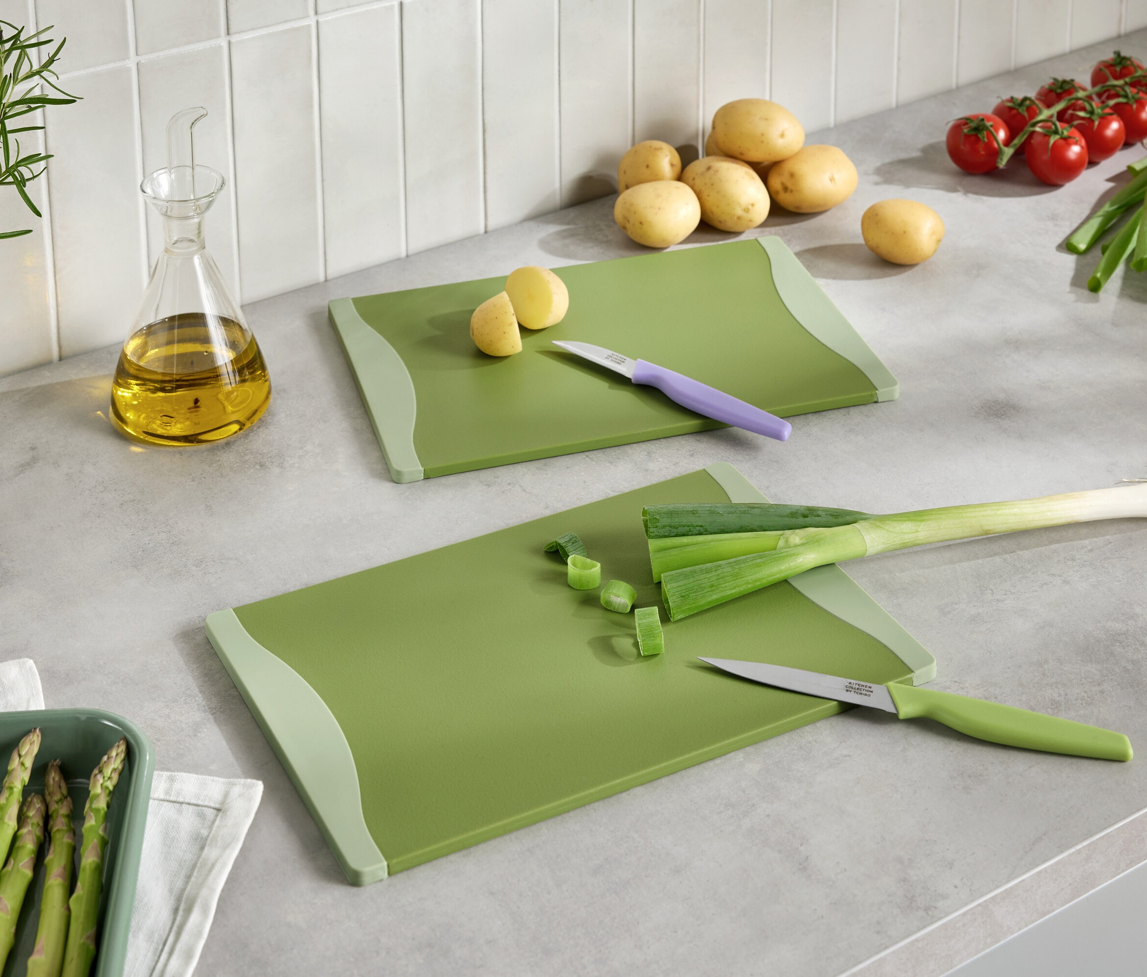 Dve zelené dosky na krájanie s nakrájanou zeleninou, nože, zemiaky, paradajky, pór, špargľa a olej na kuchynskej linke.
