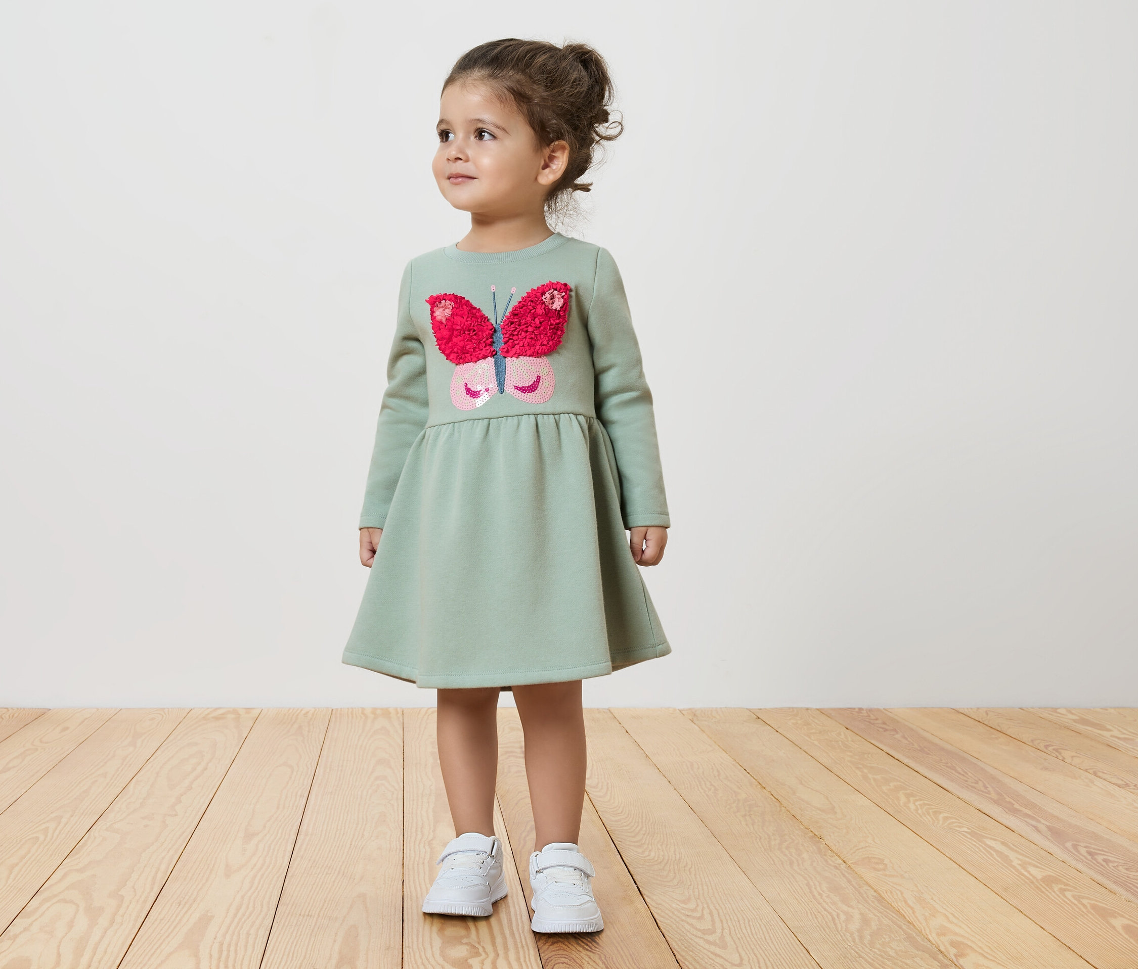 Dievča v zelených šatách s motýľom stojí na drevenej podlahe.