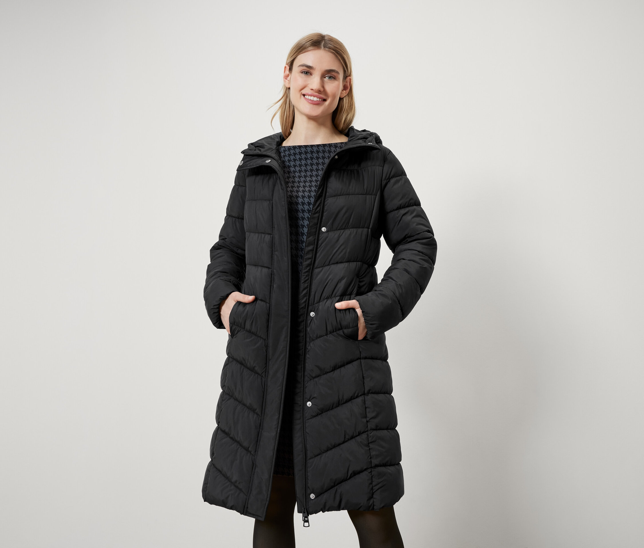Žena s blond vlasmi stojí a má na sebe dlhú čiernu prešívanú bundu s kapucňou.