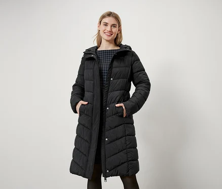 Žena s blond vlasmi stojí a má na sebe dlhú čiernu prešívanú bundu s kapucňou.