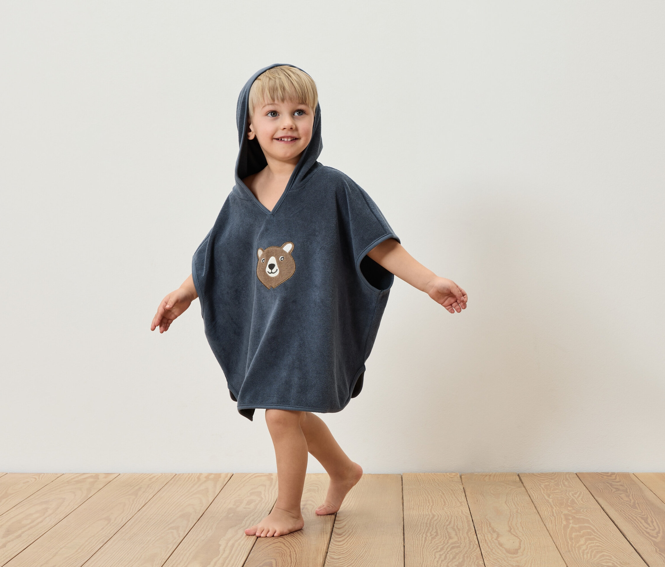 Chlapec v modrom ponču s kapucňou a medvedí aplikáciou stojí na drevenej podlahe pred bielou stenou.