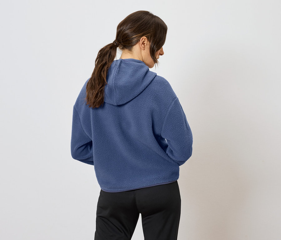 Zozadu je vidieť žena s hnedými vlasmi stiahnutými do copu, má na sebe modrú mikinu s kapucňou a čierne nohavice.