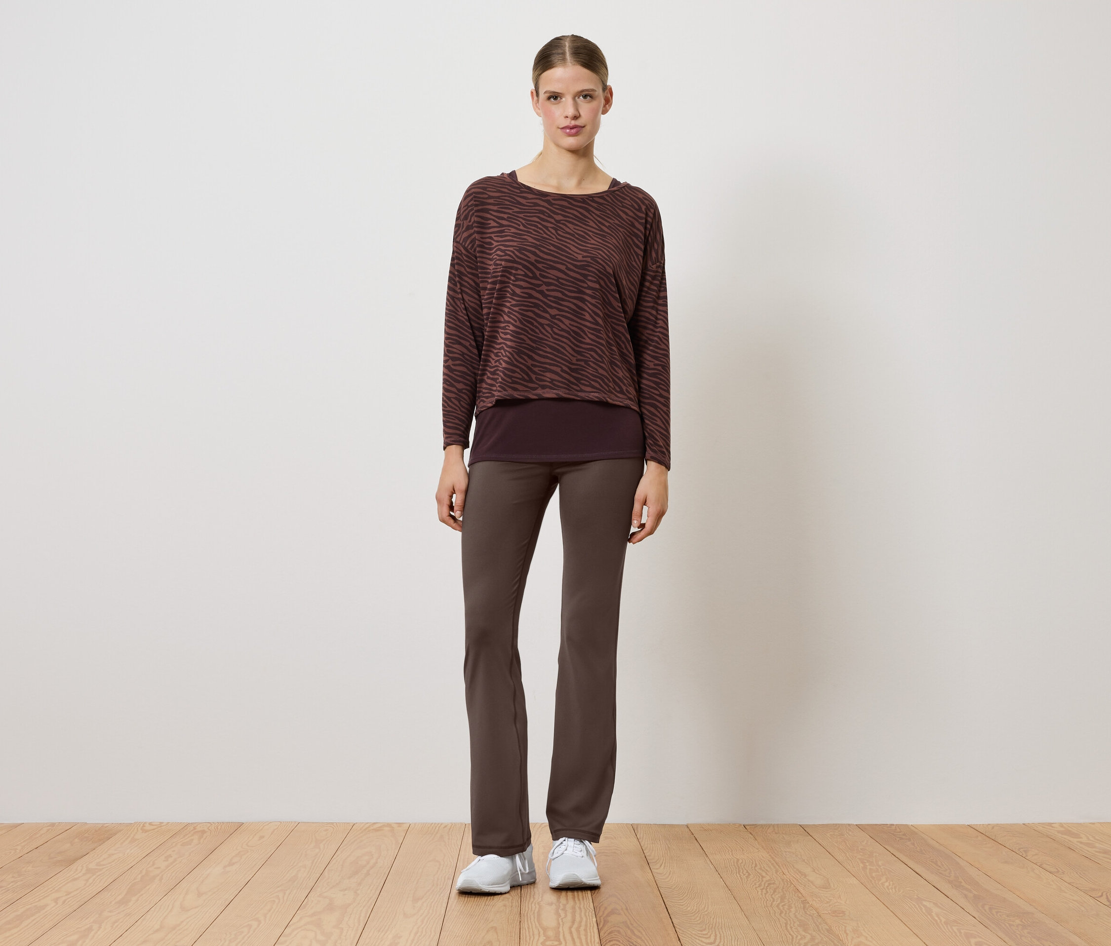 Žena v hnedom svetri a nohaviciach stojí na drevenej podlahe.