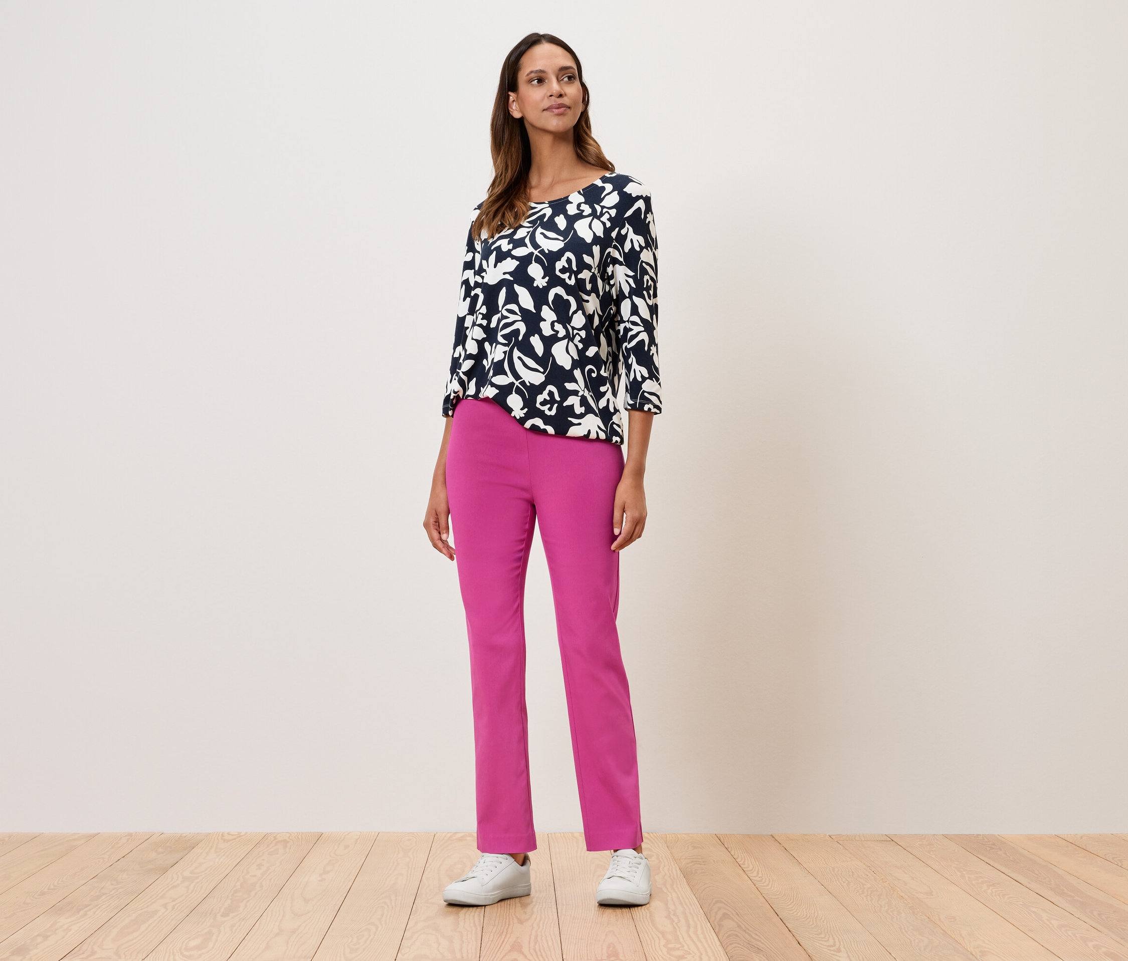 Žena stojí a má na sebe kvetinový top s trojštvrťovými rukávmi, ružové nohavice a biele tenisky.