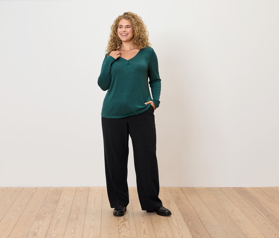 Žena s kučeravými vlasmi pózuje v smaragdovo zelenom svetri a čiernych nohaviciach.